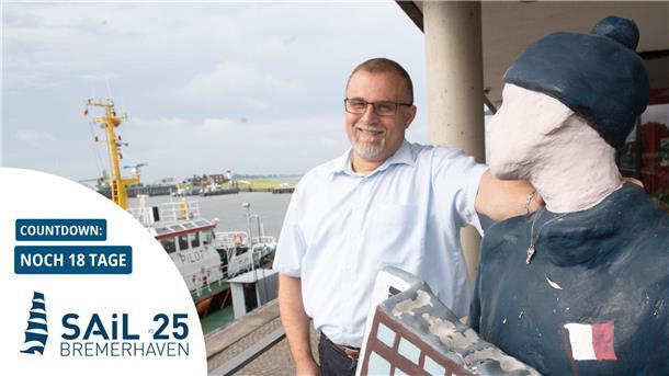 Mann steht neben einer Figur, im Hintergrund ein Schiff