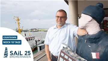 Mann steht neben einer Figur, im Hintergrund ein Schiff