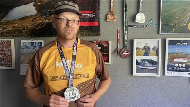Mann mit Medaille um den Hals vor einer Wand mit weiteren Urkunden und Medaillen.