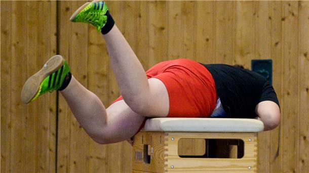 Ein übergewichtiger Junge rollt sich im Sportunterricht über einen Kasten. 