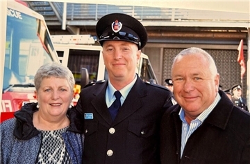 Eine Frau und zwei Männer. Der Mann in der Mitte trägt eine Uniform und einen Hut. Der Mann in der Mitte ist offenkundig ein Feuerwehrmann.
