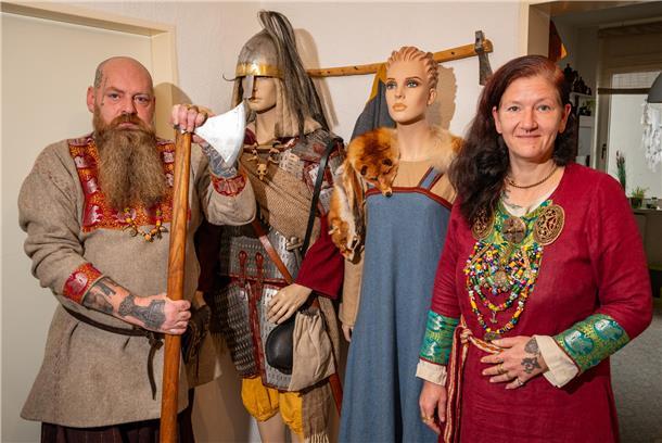 Ein Mann mit Glatze und langem Bart hat eine Axt in der Hand. Neben ihm stehen zwei Figuren. Eine trägt einen Helm und eine Tunika, die andere ein blaues Kleid. Neben den Figuren steht eine Frau mit dunkelroten Haaren. Sie hat eine weinrote Tunika an. 