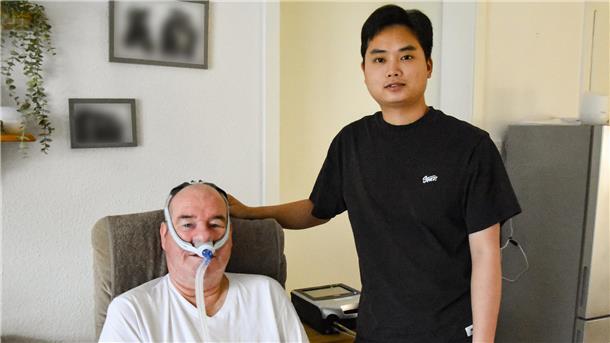 Mit neuem Pflegeteam: ALS-Patient lebt wieder in Zeven Ein älterer Mann mit Maskenbeatmung sitzt lächelnd auf einem Sessel, neben ihm steht ein junger Mann.