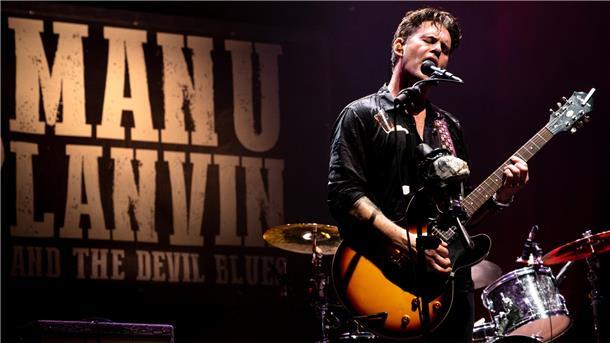 Musiker Manu Lanvin steht auf einer Bühne, spielt E-Gitarre und singt leidenschaftlich ins Mikrofon. Im Hintergrund ist ein großes Banner mit der Aufschrift „MANU LANVIN AND THE DEVIL BLUES“ zu sehen.