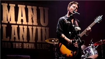 Musiker Manu Lanvin steht auf einer Bühne, spielt E-Gitarre und singt leidenschaftlich ins Mikrofon. Im Hintergrund ist ein großes Banner mit der Aufschrift „MANU LANVIN AND THE DEVIL BLUES“ zu sehen.