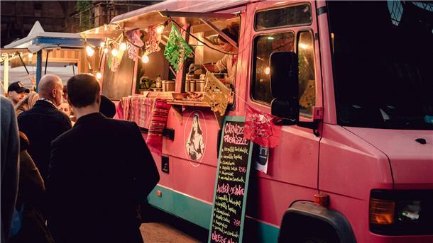 Ein rosa Foodtruck, aus dem eine Frau Essen verkauft. Davor stehen viele Gäste und warten auf ihr Essen.