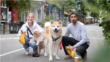Maxima und Hussein gehen mit ihren Hunden durch die Sommerstraße. Kaffee und Brötchen fürs Frühstück haben sie dabei.