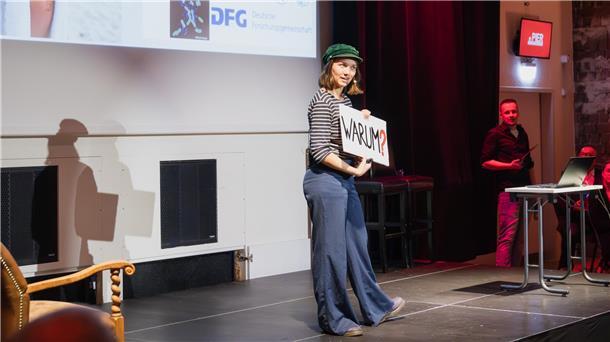„Science goes public“ startet mit einem „süßen Thema“ Eine Person steht auf einer Bühne, hält ein Schild mit der Aufschrift „WARUM?“ und spricht zu einem Publikum. Im Hintergrund sind eine Leinwand mit Logos und ein Moderator am Rand der Bühne zu sehen.