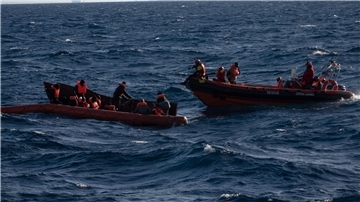 Mehrere Migranten sitzen in einem Boot im Mittelmeer während Rettungskräfte versuchen ihnen zu helfen.