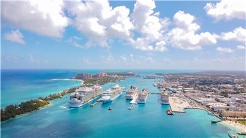 Luftaufnahme des Kreuzfahrthafens von Nassau auf den Bahamas: Mehrere große Kreuzfahrtschiffe liegen nebeneinander an den Piers. Im Hintergrund erstreckt sich die Stadt mit Hotels und Gebäuden, links das türkisfarbene Meer und Sandstrände.