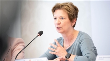 Eine Frau spricht in ein Mikrofon während einer Pressekonferenz oder Veranstaltung. Sie trägt ein hellblaues Oberteil, eine Smartwatch und gestikuliert mit der linken Hand. Vor ihr steht ein Namensschild auf dem Tisch.