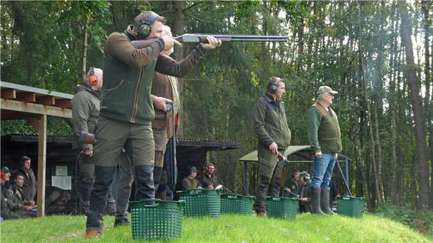 Hegering Hagen trainiert für die herbstliche Jagdsaison Mehrere Jäger stehen auf einem Schießstand im Grünen, tragen Gehörschutz und Jagdbekleidung. Einer von ihnen zielt mit einer Flinte auf eine fliegende Wurfscheibe, während andere warten. Im Hintergrund beobachten weitere Personen das Geschehen.