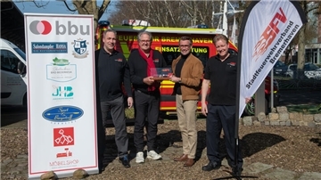 Vier Männer stehen bei Sonnenschein vor einem Feuerwehrfahrzeug und präsentieren eine Auszeichnung. Links steht ein Roll-Up mit Logos der BBU, rechts ein Banner des Stadtfeuerwehrverbands Bremerhaven.