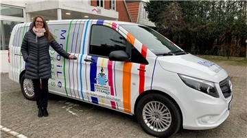 Eine Frau mit langer, dunkler Jacke und Schal steht neben einem bunt gestalteten Fahrzeug, das die Aufschrift „MobiLe Landkreis“ trägt.