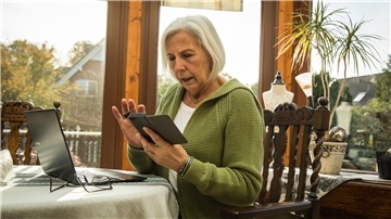 Eine ältere Frau sitzt am Tisch und hält ein Smartphone in der Hand. Vor ihr auf dem Tisch steht ein Laptop.