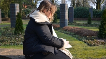 Eine junge Frau sitzt auf einem Friedhof und schreibt einen Text auf einen Block. 