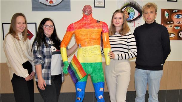 Eine in Regenbogenfarben bemalte Figur ohne Kopf steht in der Mitte. Links und rechts davon stehen drei Schülerinnen und ein Schüler der Waldschule Hagen-Beverstedt.