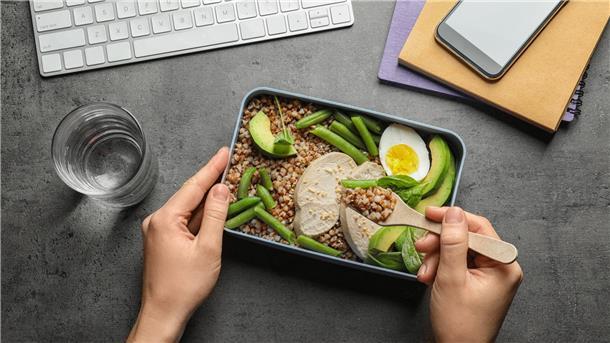 Eine Meal Prep Box mit Bohnen, Ei, Getreide, Avocado und Tofu stehen auf einem grauen Tisch, auf dem auch ein Glas Wasser, eine weiße Tastatur und Hefte liegen.