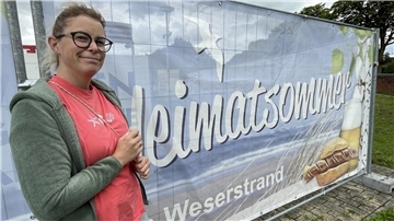 Eine Frau steht neben einem Plakat. Mitzi Brunke veranstaltet zusammen mit ihrem Mann Klaus den Heimatsommer.