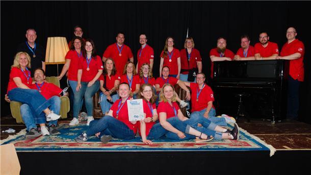 Mit Musik und Teamgeist: TuSpo Surheide feiert Erfolg in Leipzig Das Turnermusiker-Orchester des TuSpo Surheide posiert in roten Shirts mit Medaillen und Urkunde auf einer Bühne vor schwarzem Hintergrund, umgeben von Sofa, Sessel, Teppich und Klavier.