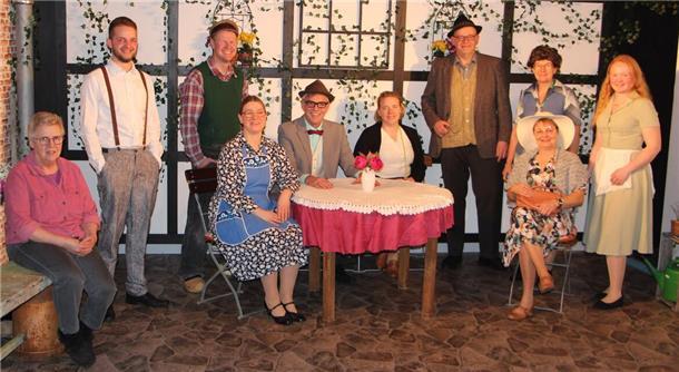 Eine Gruppe von zehn Personen in traditionell oder retro anmutender Kleidung posiert auf einer Theaterbühne mit ländlichem oder dörflichem Hintergrund. Die Kulisse zeigt eine mit Efeu bewachsene Fachwerkwand, ein Tisch mit Spitzentischdecke und Blumen sowie rustikale Möbel.