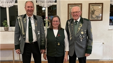 Drei Schützen in traditioneller Uniform stehen nebeneinander und lächeln in die Kamera. Zwei Männer tragen graue Schützenjacken mit Orden, die Frau in der Mitte eine grüne Weste mit Anstecknadel. Im Hintergrund sind Fenster und gerahmte Urkunden zu sehen.