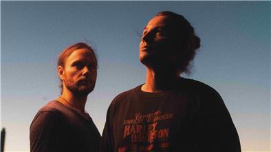 Zwei in Sonnenschein getauchte Männer stehen vor einem blauen Himmel. 