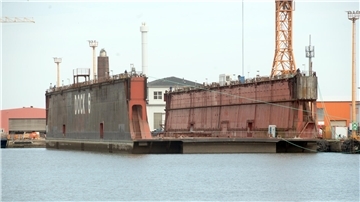 Das über 60 Jahre alte Schwimmdock 6 liegt vertäut im Hafen von Bremerhaven. Auf der Seite ist in großen weißen Buchstaben „DOCK 6“ zu lesen. Im Hintergrund sind Werftgebäude und ein orangefarbener Kran zu sehen.