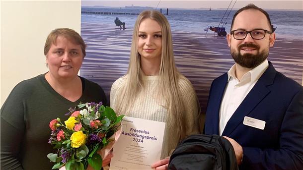 Helios-Klinik Wesermarsch: Darum ist eine Auszubildende jetzt geehrt worden Zwei Frauen und ein Mann stehen vor einer Wand. Sie halten Blumen, eine Urkunde und einen Rucksack in den Händen.
