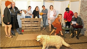Frauen und Männer proben auf einer Bühne. Durch das Bild läuft ein Hund.