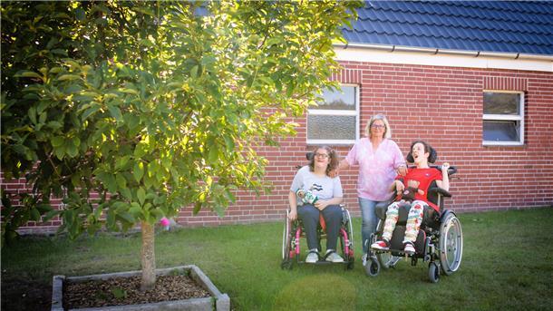 Zwei junge Frauen mit Behinderung stehen mit Ihrem Rollstuhl im Garten vor einer Backsteinwand, ihre Mutter steht zwischen ihnen. Lins im Vordergrund wächst ein Baum. 
