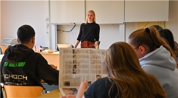 Eine Frau steht in einem Klassenzimmer an einer Tafel, Schüler sitzen im Klassenraum und hören zu.