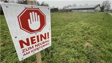 Ein Schild am Rande einer Grünfläche. Im Hintergrund ist eine Halle zu sehen.