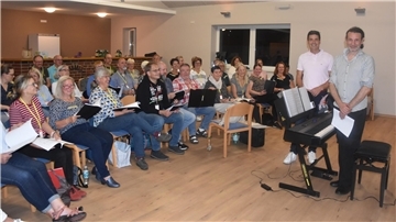 Etwa 25 Menschen sitzen im Halbkreis auf Stuhlreihen und halten Schnellhefter in der Hand. Davor stehen zwei Männer neben einem elektrischen Piano. 
