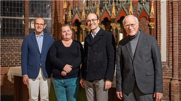 Drei ältere Männer und eine Frau in einer Kirche blicken in die Kamera. 