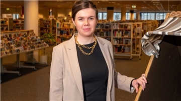 Eine Frau steht in der öffentlichen Bibliothek, im Hintergrund sind Regale voller Bücher zu sehen.