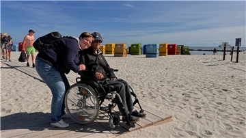 An einem Strand steht eine Frau mit einem Mann im Rollstuhl. Beide haben ein Lächeln im Gesicht.