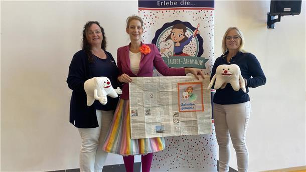 Drei Frauen stehen nebeneinander und präsentieren Zahn-Stofftiere sowie eine aufgeschlagene Zeitung. 