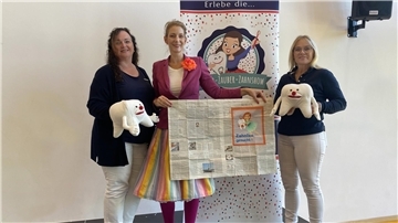 Drei Frauen stehen nebeneinander und präsentieren Zahn-Stofftiere sowie eine aufgeschlagene Zeitung. 