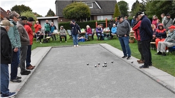 Mehrere Personen stehen um ein Boule-Spielfeld, während ein Spieler eine Kugel wirft. Zuschauer sitzen auf Bänken am Spielfeldrand, im Hintergrund sind Wohnhäuser zu sehen.