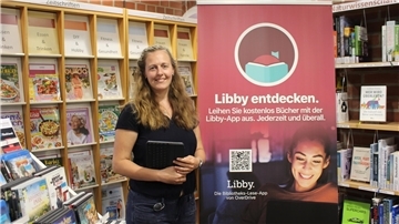Frau hält ein Tablet in der Hand und steht neben einem Plakat auf dem geschrieben ist: "Libby entdecken" 