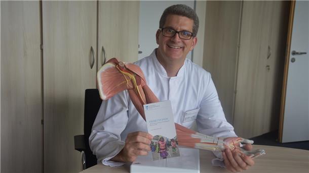 Ein Arzt im weißen Kittel lächelt in die Kamera. Vor sich auf einem Tisch hält er einen Arm aus Kunststoff, bei dem Muskeln und Blutgefäße dargestellt sind.