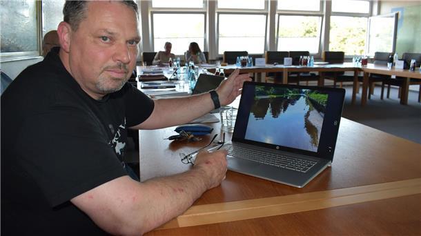 Mann guckt in die Kamera und zeigt gleichzeitig ein Überflutungsfoto auf einem Laptop. 