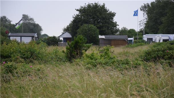 Holzbuden und Wohnwagen im Hintergrund, Gras und Büsche im Vordergrund