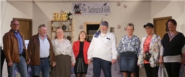 Die plattdeutsche Theatergruppe „Lüüd von Ossentorm“ steht nach der Aufführung gemeinsam auf der Bühne und hält sich an den Händen. Im Hintergrund ist das Bühnenbild mit einem Bar-Regal und einem Schild mit der Aufschrift „De Satansköök“ zu sehen.