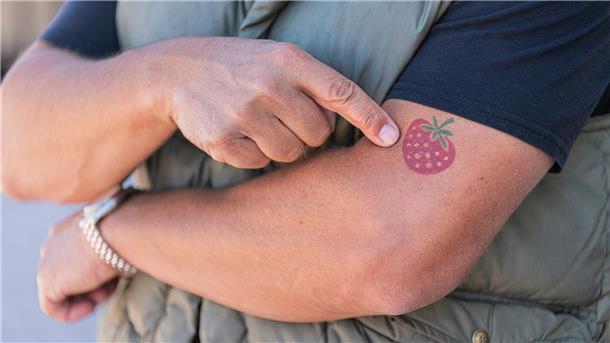 Ein Mann zeigt auf ein Erdbeer-Tattoo auf seinem linken Arm.