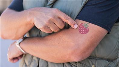 Ein Mann zeigt auf ein Erdbeer-Tattoo auf seinem linken Arm.