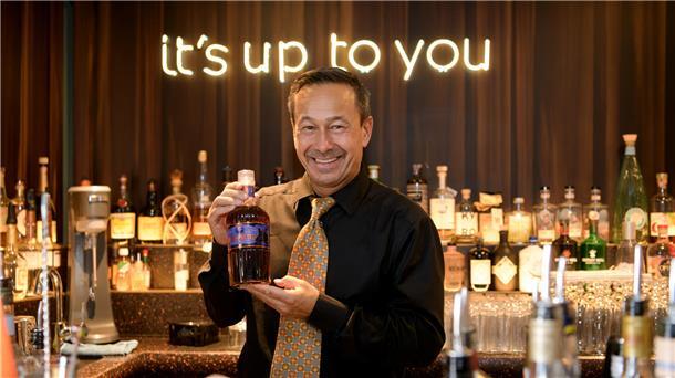 Barkeeper Ron de Preter bringt seine erste eigene Rumkreation heraus Ein Mann in einer Bar, der seine Rumkreation zeigt. Im Hintergrund ist in leuchtender Schrift "it's up to you" zu lesen.