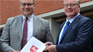Zwei Männer halten ein Papier mit dem aufgedruckten Wappen Niedersachsens in Händen. 