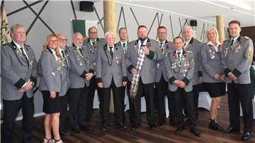 Gruppenfoto von Mitgliedern eines Schützenvereins in festlicher Uniform mit Orden und Abzeichen. In der Mitte steht der Schützenkönig mit der Schärpe. Die Personen posieren in einem Saal mit Schützenfahne und dekorativer Wand im Hintergrund.